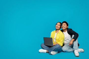 Smiling Asian couple in casual clothes sitting with a laptop, looking up with hopeful expressions