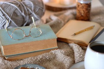 Fototapeta premium Cup of tea or coffee, cookies, books, glasses, e-reader, almonds, ball of yarn, knitting needles and lit candles on the table. Hygge at home. Selective focus.