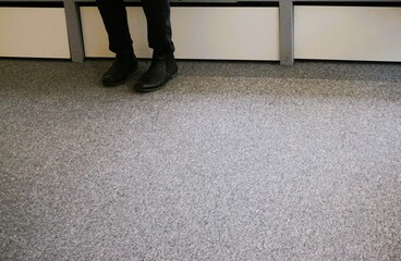 Schwarze Lederschuhe und schwarze Jeanshosenbeine von Mann auf grauem Teppichboden vor weißen...