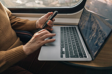 Hands of unrecognizable woman holding smartphone next to laptop while traveling in train