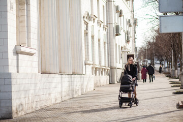 A young mother iswalking around the city with a black stroller and a small child. Woman is carrying a black carriage