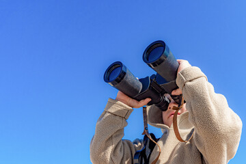 A boy with binoculars on a background of blue sky looks up.