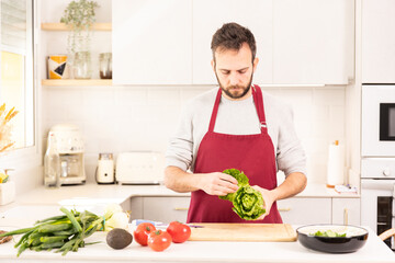 A man is preparing a salad in a kitchen. He is wearing a red apron and holding a head of lettuce. The kitchen is well-equipped with appliances such as a toaster, microwave, and oven