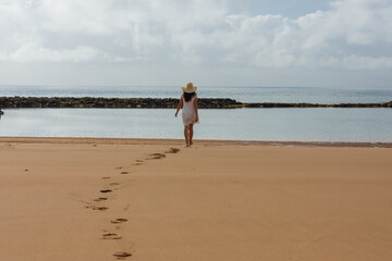 Woman with a hat walking towards the beach leaving footprints in the sand