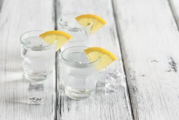 Cold vodka in shot glasses with lemon slices and ice cubes on a white wooden table. Side view, selective focus. - 762496902