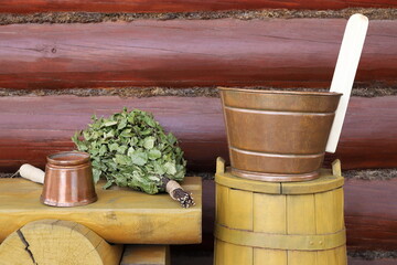 On the yellow wooden bench and barrel against the log wooden background there are copper sauna accessories and dry birch broom. 