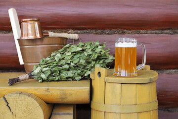 A glass mug of beer is on the wooden barrel next to copper sauna accessories and dry birch broom that are on the yellow wooden bench.