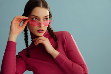 Fashionable confident woman wearing trendy rimless pink sunglasses, turtleneck, posing on blue background. Studio fashion portrait. Copy, empty, blank space for text