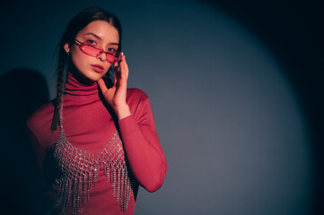 Fashionable confident woman wearing trendy pink sunglasses, turtleneck, rhinestone top, posing on dark background. Studio fashion portrait. Copy, empty, blank space for text