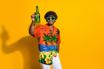 Portrait of handsome guy with afro hair wear hawaii shirt in glasses raising bottle of beer say cheers isolated on yellow color background