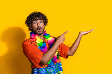 Photo of astonished man dressed print shirt flower necklace directing at offer on arm empty space isolated on yellow color background