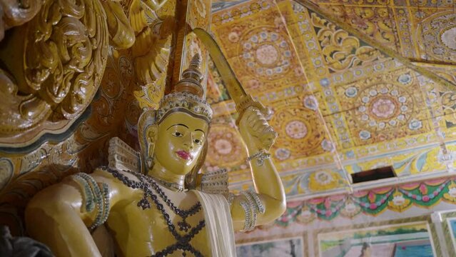 Golden Buddha statue stands in ornate temple. Traditional art adorns walls, ceiling with vibrant colors, patterns. Sacred space filled with cultural, religious symbols for meditation, peace.