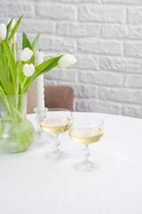 Table set with two glasses of white wine, vase with white tulips, candle on white table on bricks background.