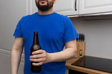 Mężczyzna trzymający butelkę piwa, ubrany w niebieską koszulkę, koncepcja problemów związanych z nadmiernym spożyciem alkoholu w społeczeństwie. - obrazy, fototapety, plakaty