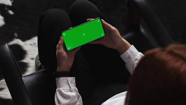 Close-up woman hands watching on the horizontal green screen phone sitting in a chair. Green screen cromakey phone used by woman . Technology lifestyle.