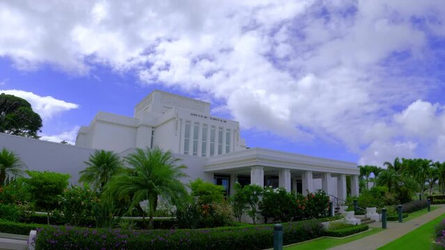 Side view of Laie Hawaii Temple surrounded by flowers and greenery. Establishing shot of white Mormon church on a cloudy day.
