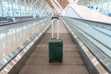 Green stylish suitcase on travelator against a sparkling light empty blurred airport background. Concept of business travel, summer vacation, adventure