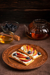 Homemade sandwich made with crispbread decorated with cream cheese, sliced nectarine fruit, blueberries and seeds served on brown plate on table with glass tea pot and honey in bowl for breakfast