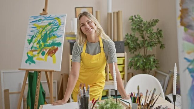 A young, blonde woman artist in a bright yellow apron stands in an art studio with a colorful painting on an easel.