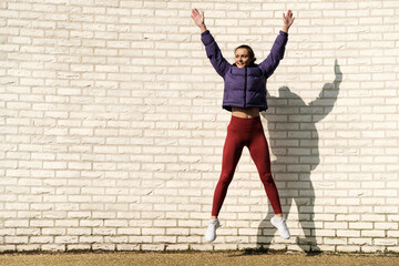 Energetic sport woman jumping in front of white brick wall outdoors.