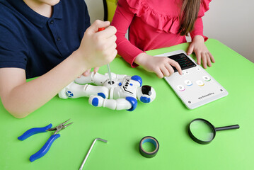 Children's hands assemble robot on workbench, screwing parts while reviewing programming code on tablet. Concept of learning and hands-on education