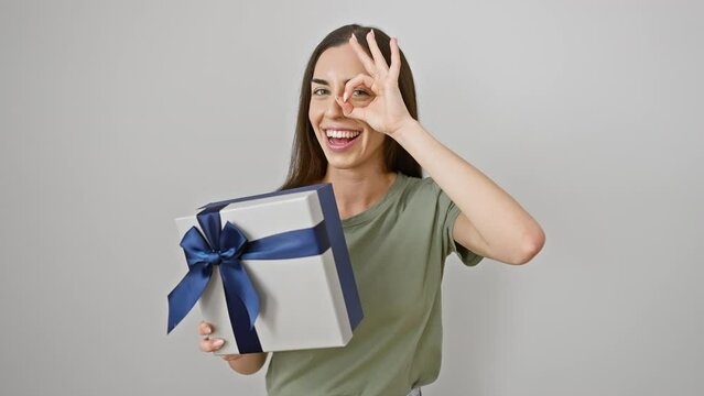 Joyful young hispanic woman makes a fun 'ok' gesture, peering through her fingers, her smile beaming as she holds her beautifully wrapped birthday gift, all against an isolated white background.