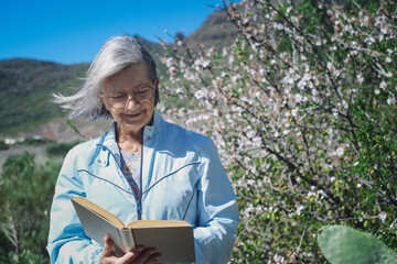 Smiling senior woman with white hair sitting outdoors in sunny day near flowering almond trees...