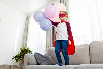 Boy in superhero costume with balloons standing on sofa