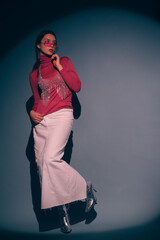 Fashionable confident woman wearing trendy pink sunglasses, turtleneck, rhinestone top, white maxi skirt, silver ankle boots, posing on dark background. Full-length studio fashion portrait