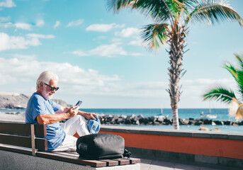 Senior bearded man using smartphone sitting outdoors on bench face the sea enjoying sunny day, travel, vacation, retirement lifestyle