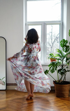 attractive woman with graceful forms in a beautiful dress with a floral print is dancing in a spacious apartment against the background of a bright window