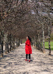 Fototapeta na wymiar Portrait of a stylish woman outdoors. A model with dark hair, in a red raincoat and red high-heeled shoes walks along an alley among the trees in the park. Turns around and looks at the camera