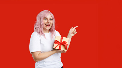 Pretty cheerful woman with pink hair holding present and showing finger away at copy space isolated over red background. Girl holding wrapped present, celebrating birthday. Special offer