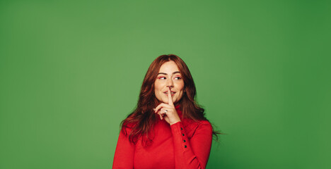 Fashionable woman standing on vibrant green background, quietly gesturing shush with finger