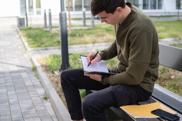 A young male student holding a notebook and sitting on a bench in front of a building, taking notes and learning for the exam on a sunny day