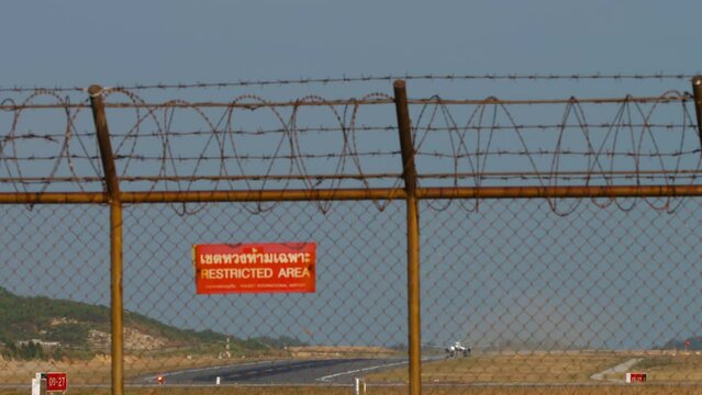 Airport fence with red Restricted Area sign. Jet plane with an unrecognizable livery taking off and climbing, front view, long shot