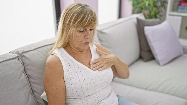 At home on her cozy sofa, a beautiful blonde middle-aged woman suffering a heart attack, hands clutching chest in despair, despite the warm sunlight filtering through the living room
