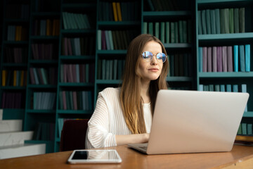 Focused young woman with sunglasses working on laptop in a library with colorful bookshelves.