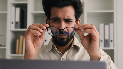 Unhealthy Indian Arabian man in glasses for laptop business work confused businessman office ceo with bad eyesight blurry vision eyes myopia take off spectacles medical eye laser correction loss sight