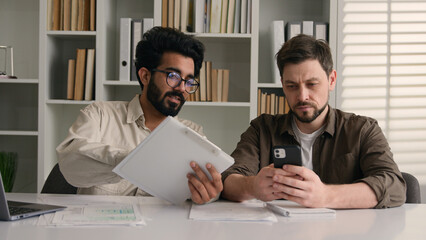 Two businessmen multiracial men co-workers in office Arabian Indian businessman show papers business project on laptop to colleague Caucasian man ignoring addicted mobile phone inefficient teamwork