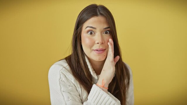 Hush! beautiful young hispanic woman wearing sweater, whispering juicy secrets. standing over isolated yellow background, hand on mouth, stirring tempting chatter conversation.