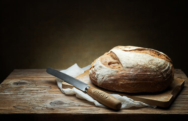 Whole loaf of traditional bread with knife, cutting board and tea towel on old wooden table, close-up, space for text.