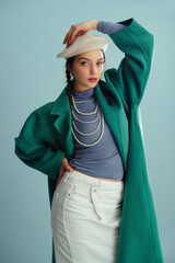 Fashionable confident woman wearing white beret, green coat, blue turtleneck, layered pearl necklace, denim skirt, posing on blue background