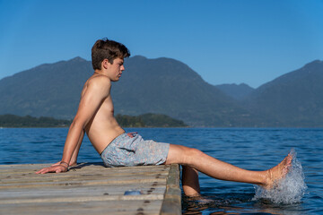 Teenage boy sitting on the lake dock, dipping his feet in the water, pondering and reflecting on life.