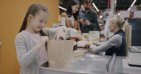 Cute little girl shopping in supermarket packing purchase in paper bag at counter with cashier. Childhood and consumerism concept.