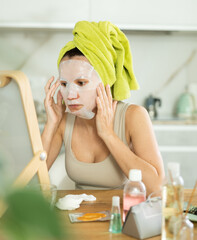 Obraz na płótnie Canvas Relaxed woman in her middle age using tissue face mask for skincare sitting at the mirror in home environment