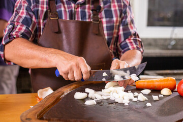 A white man wearing a leather apron and a plaid shirt slicing an onion next to a carrot on a rustic wooden board to cook dinner or lunch