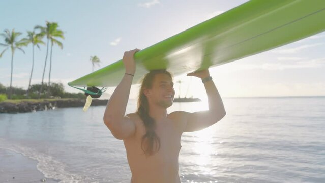 Young handsome athletic smiling man without a shirt balancing a surfboard on his head while walking along the sandy beach, the sun setting over the horizon. High quality 4k footage