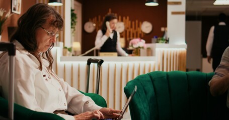 Senior couple passing time in lounge area, woman using laptop to create holiday itinerary while man enjoys lecture on couch. Retired people waiting to see accommodation at hotel. - obrazy, fototapety, plakaty