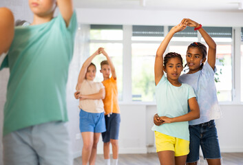 Cheerful dark-skinned preteen boy and girl training movements of slow foxtrot in dance studio with multiethnic group of kids..